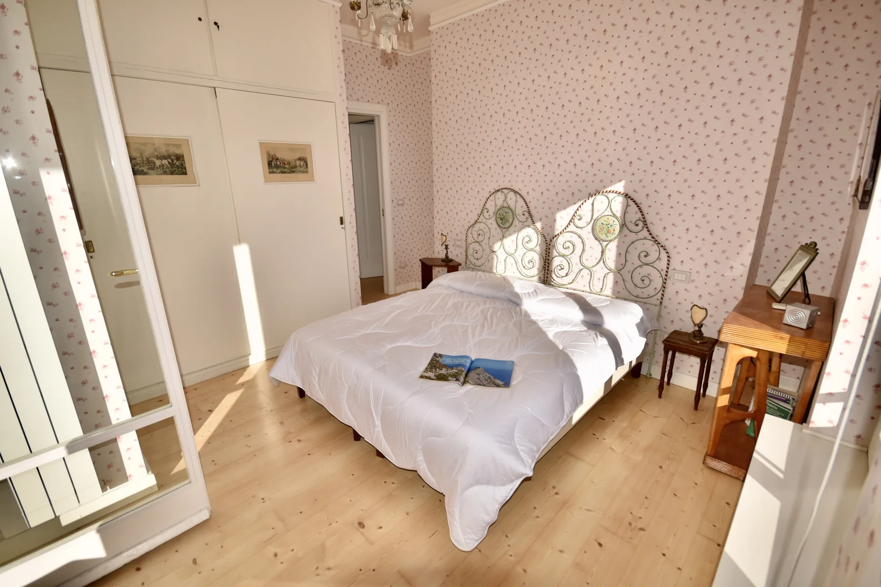 A bedroom with a white bed, patterned wallpaper, and light wood flooring.