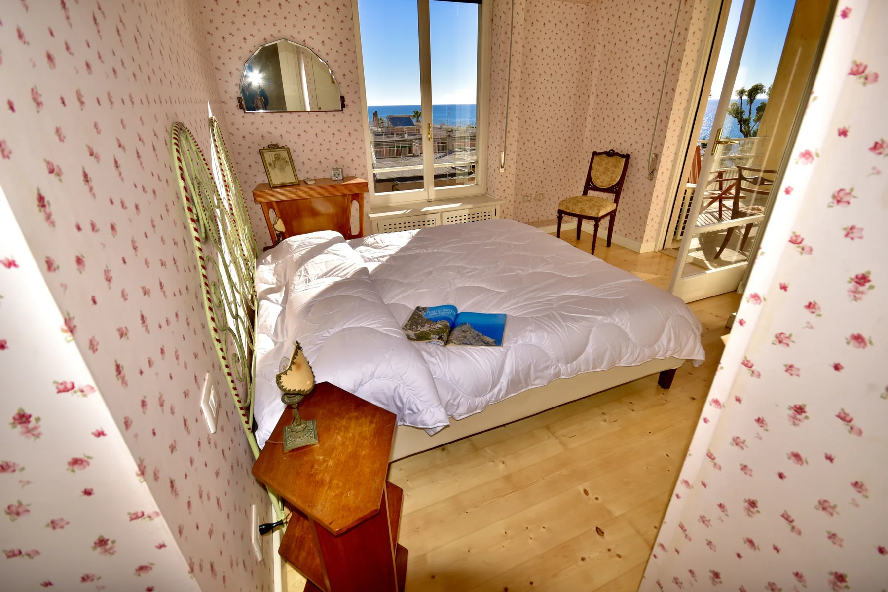 A bedroom with a bed, patterned wallpaper, and windows overlooking the sea.