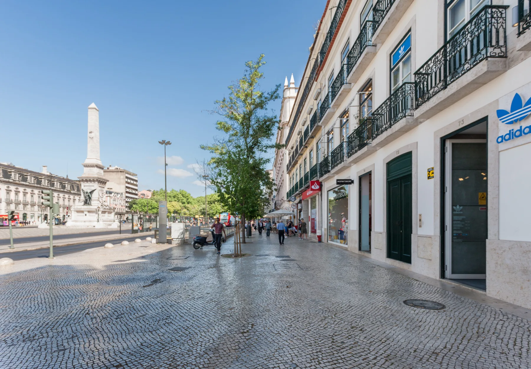 Una calle urbana con pavimento estampado, tiendas y un monumento en la plaza.