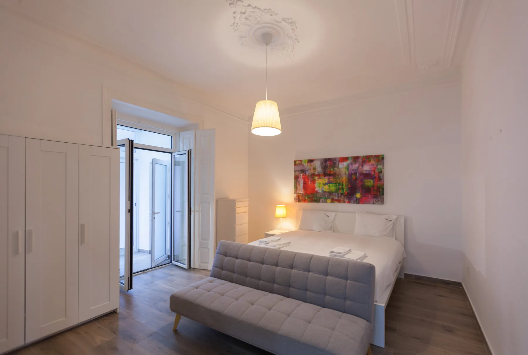 A bedroom with a bed, a gray upholstered bench, and colorful artwork above the headboard.