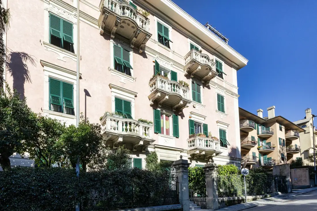 A pale multi-story building with green shutters and balconies stands behind a street-side hedge.
