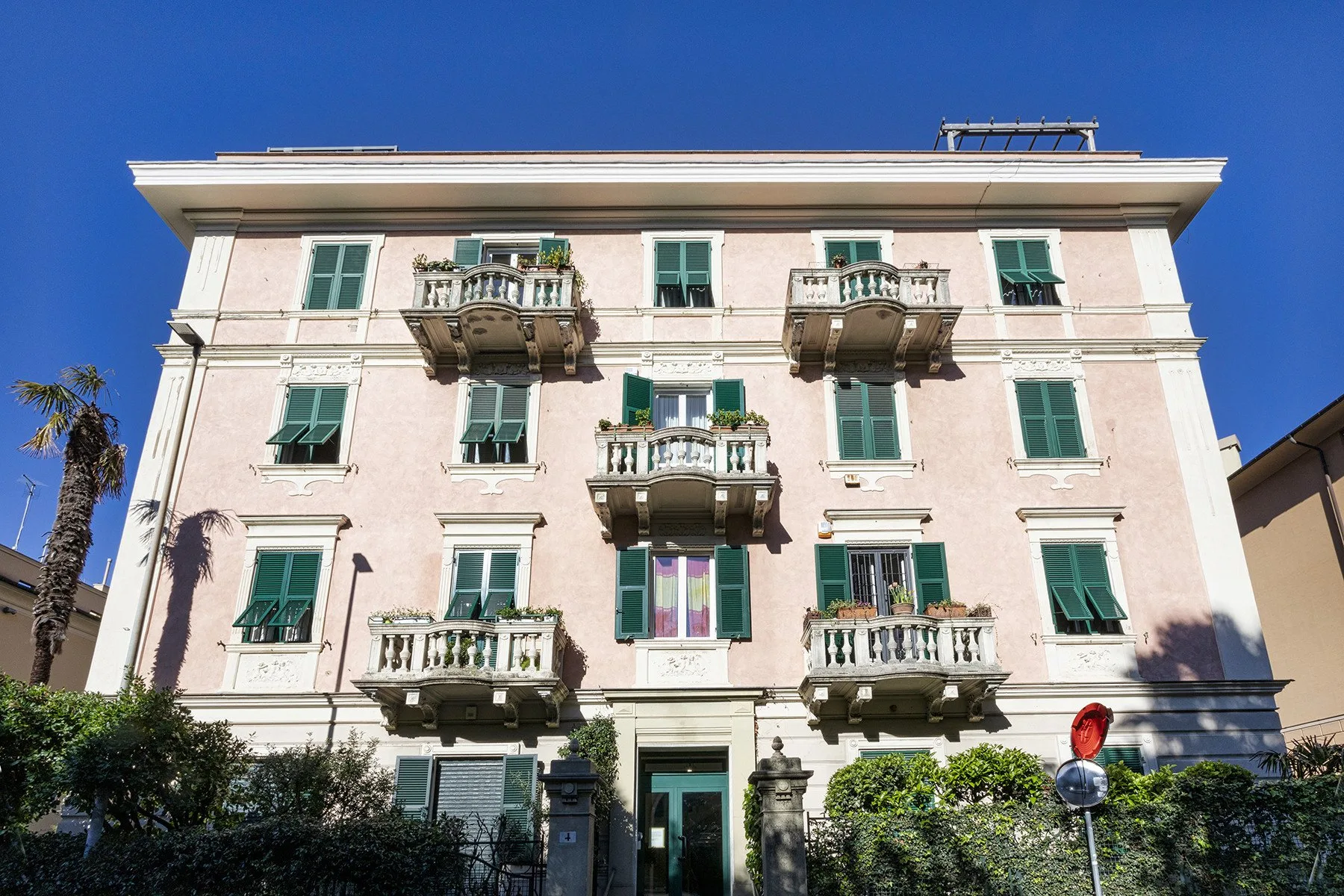 A pale multi-story building with green shutters and balconies stands against a clear blue sky.
