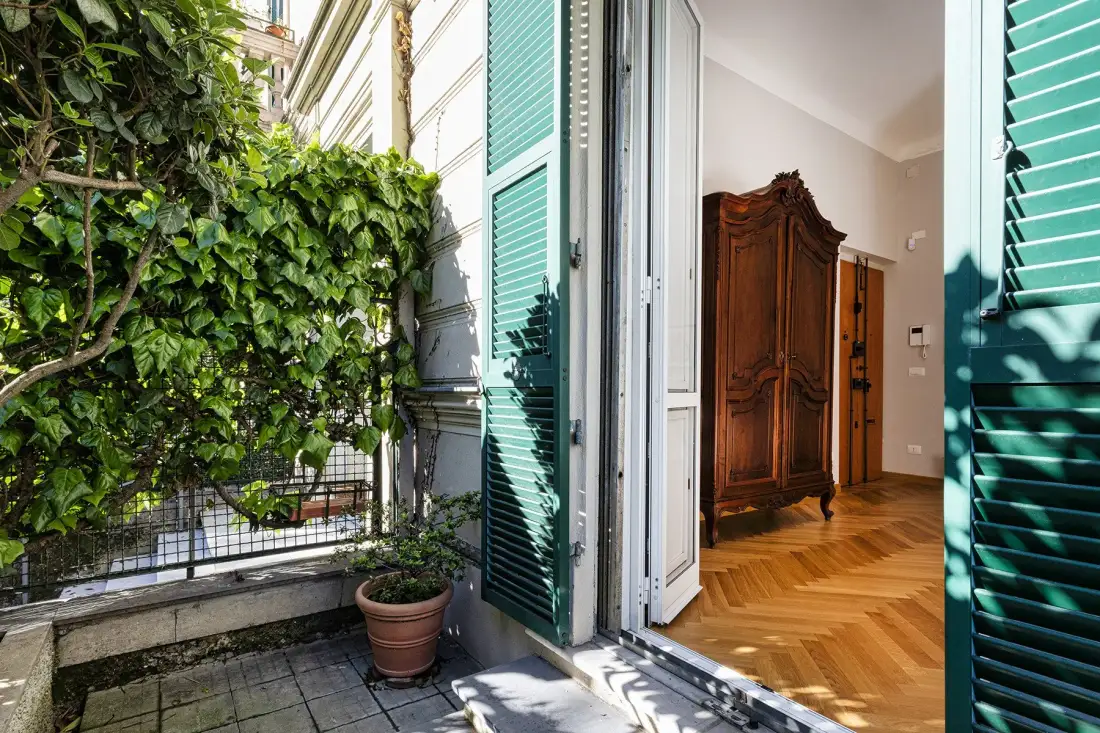 An open doorway connects a leafy patio with a room featuring a wooden wardrobe.