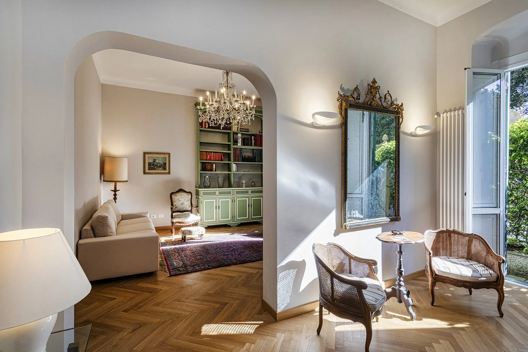 A bright sitting room with an arched opening, bookshelves, and seating by tall windows.