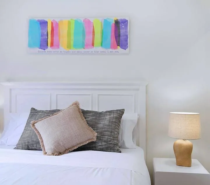 A neatly made bed with layered pillows sits beneath a colorful wall print.