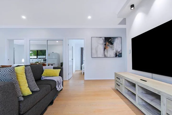 A bright living room with a sectional sofa, wall-mounted television, and light wood flooring.