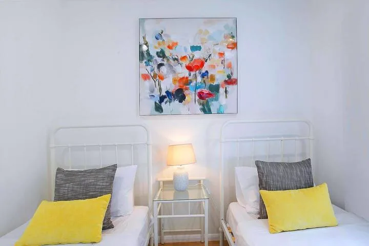 A bright bedroom with two single beds, yellow pillows, and colorful floral artwork.