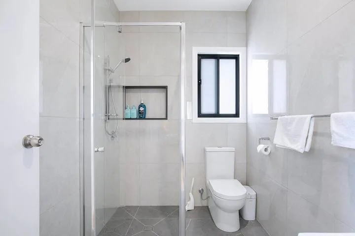 A bright tiled bathroom with a glass shower, toilet, and window.