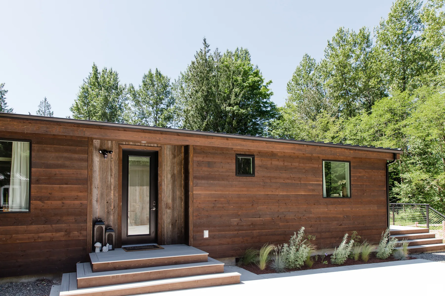 Exterior de una casa baja de madera con ventanas, escalones y árboles alrededor.