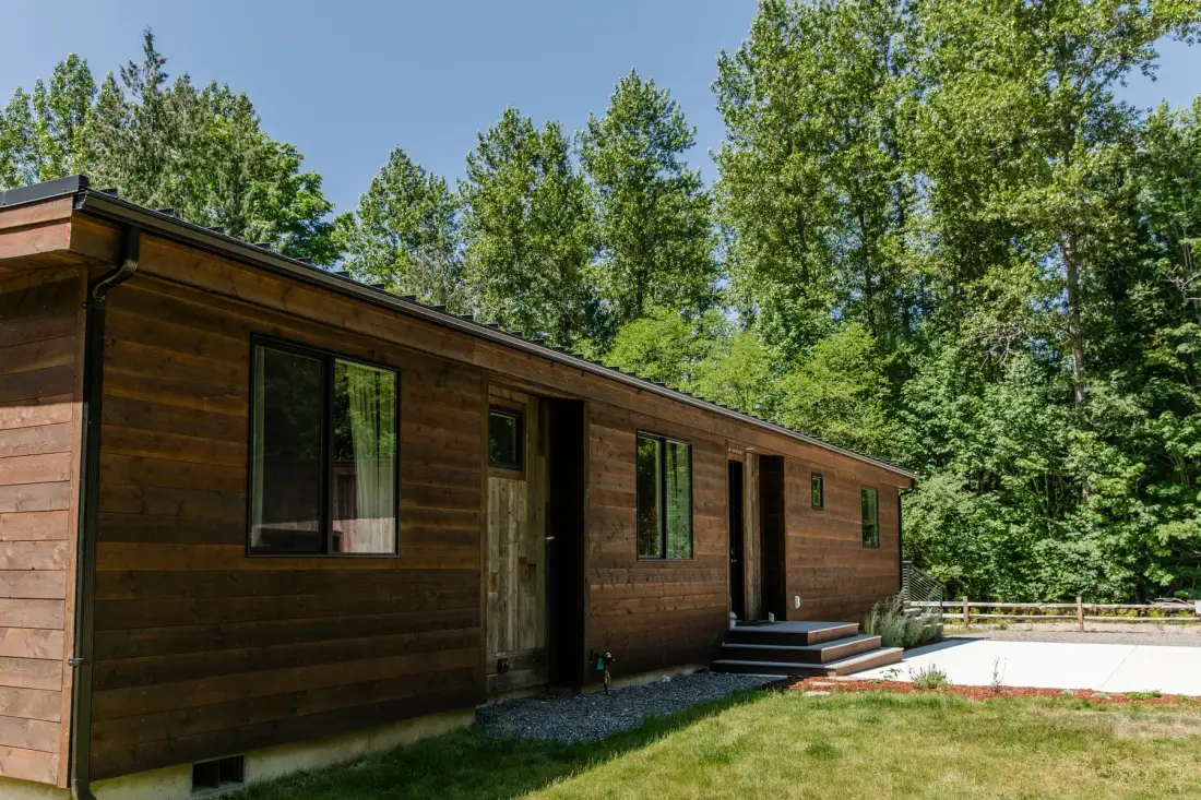 Una cabaña larga de madera se encuentra junto a un patio con césped y árboles frondosos.