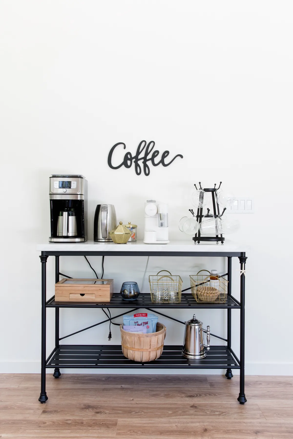 Una estación de café con cafetera y suministros está junto a una pared blanca.