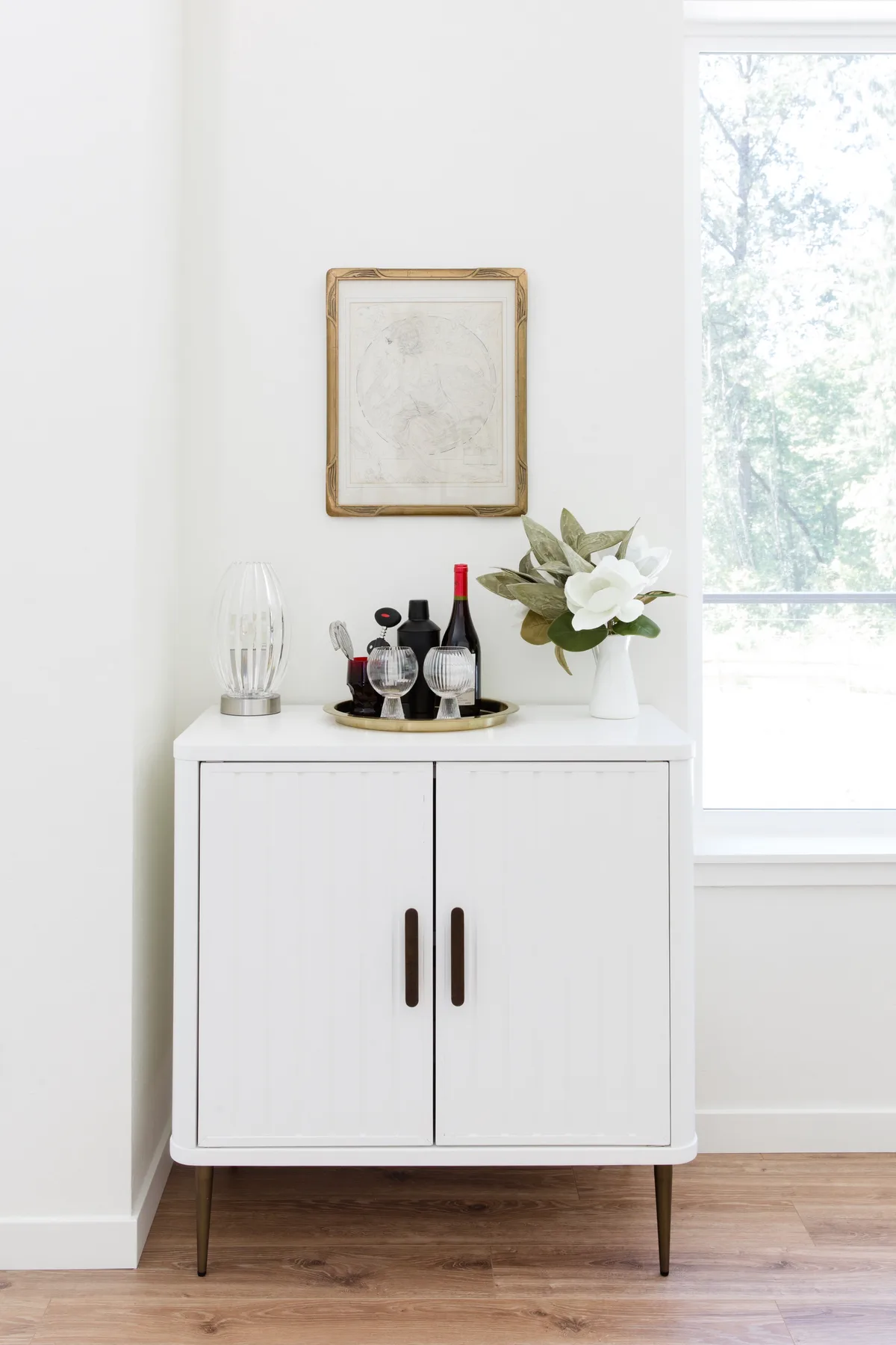 Un interior luminoso con un mueble blanco, un cuadro y flores junto a una ventana.