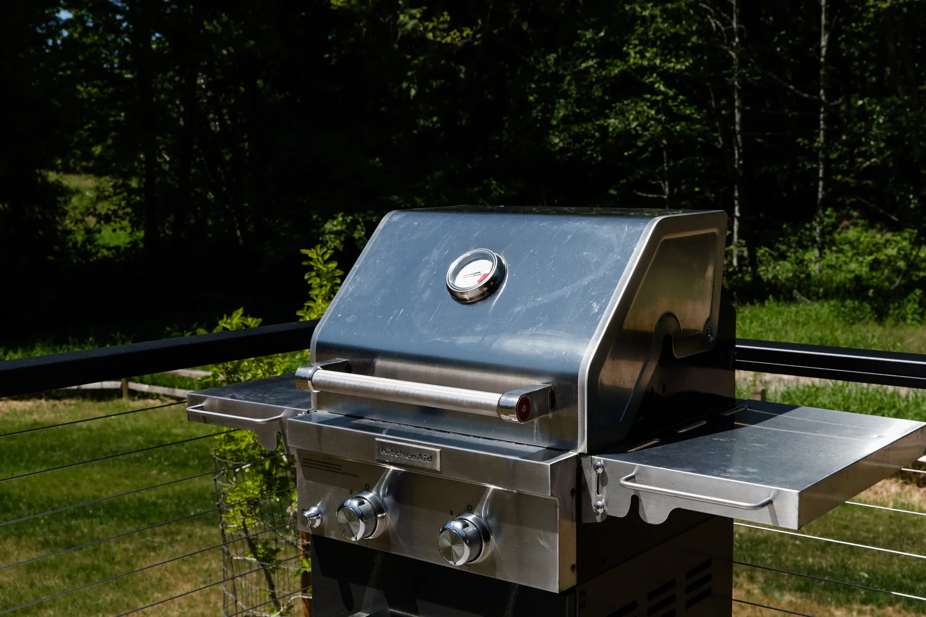 Una parrilla al aire libre con estantes laterales metálicos está junto a una zona arbolada.