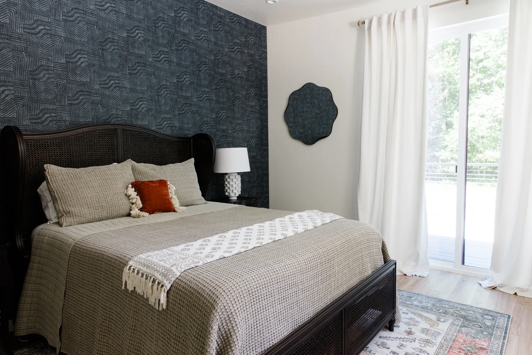Un dormitorio con una cama oscura y ropa de cama estampada junto a una ventana luminosa.