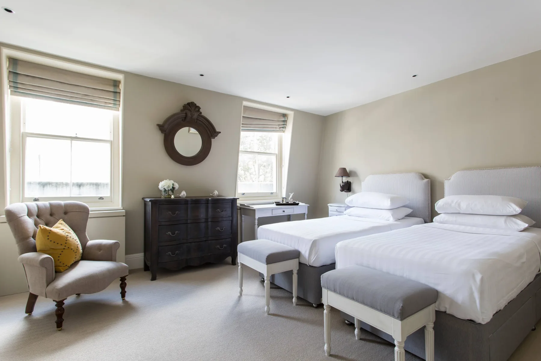 A bright bedroom with two beds, a dark dresser, and a cushioned chair.