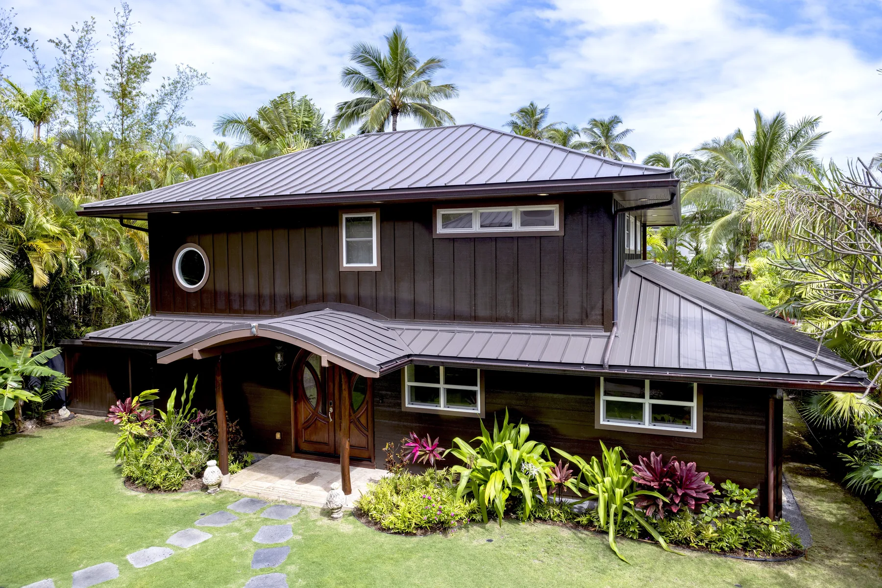 A dark wooden house with a metal roof sits among tropical plants.