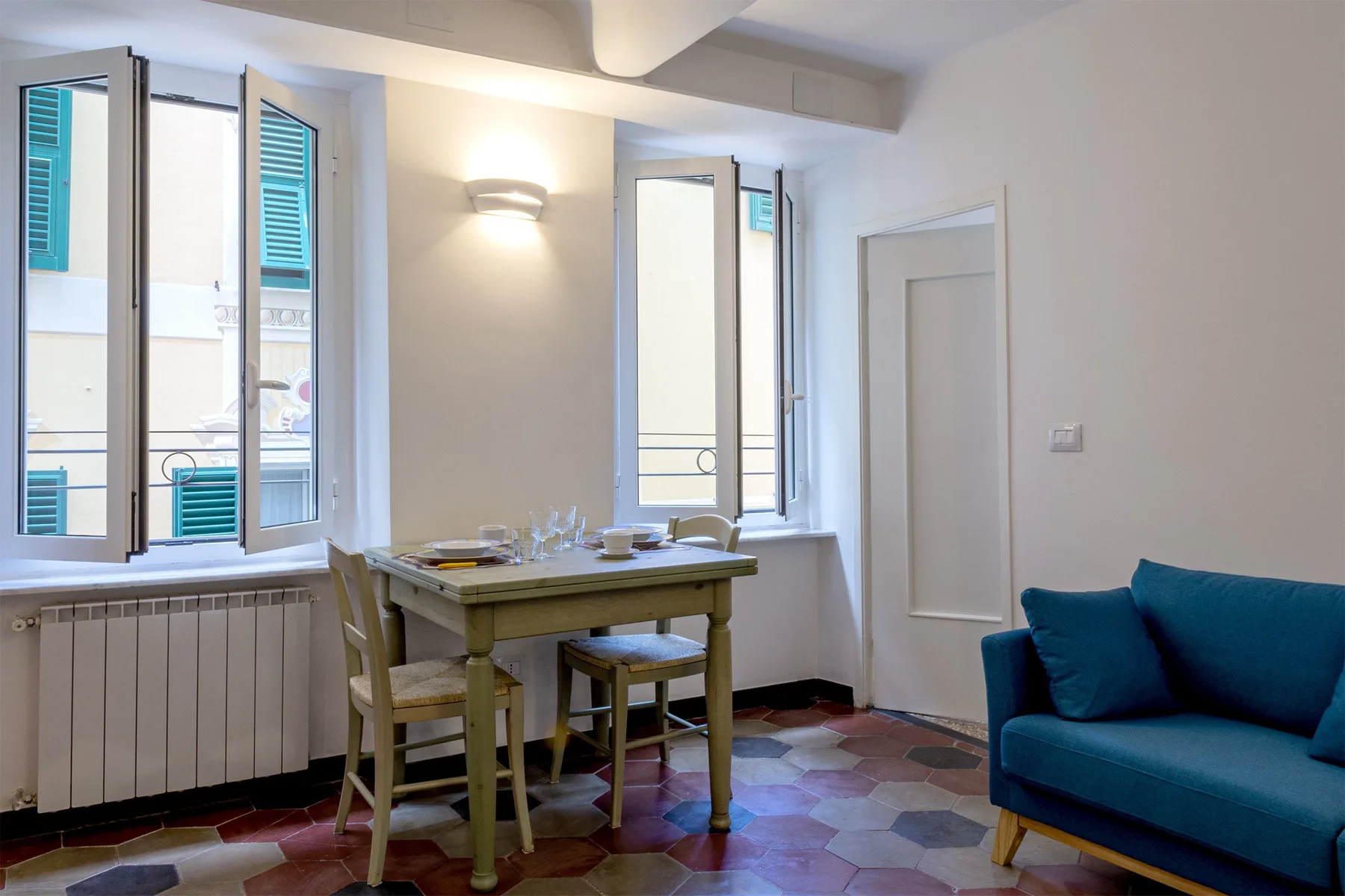 A dining area with a table and chairs beside open windows and a blue sofa.