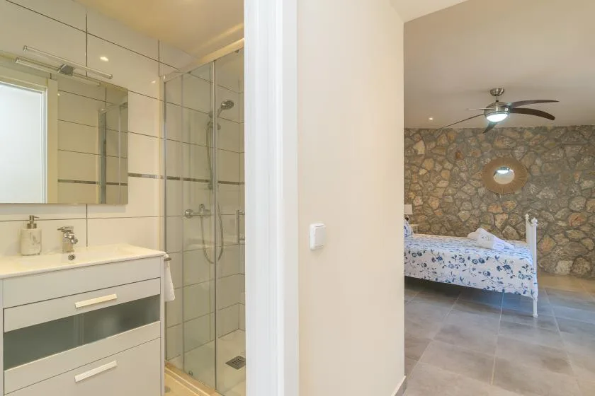 A bathroom with a glass shower is visible beside a bedroom with a stone wall.