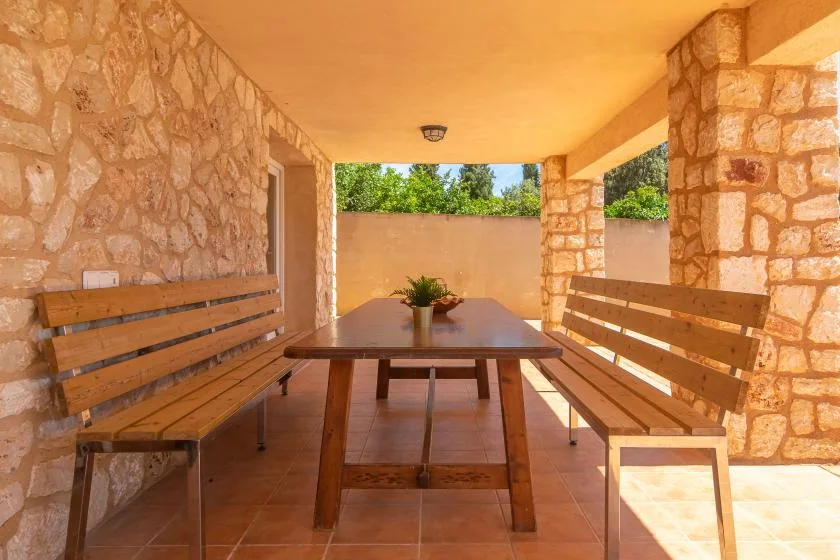 A covered patio with a wooden table and benches, open to a garden.