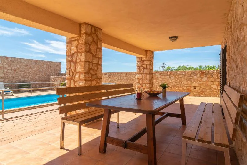 A covered stone patio with a wooden picnic table and a pool visible beyond.