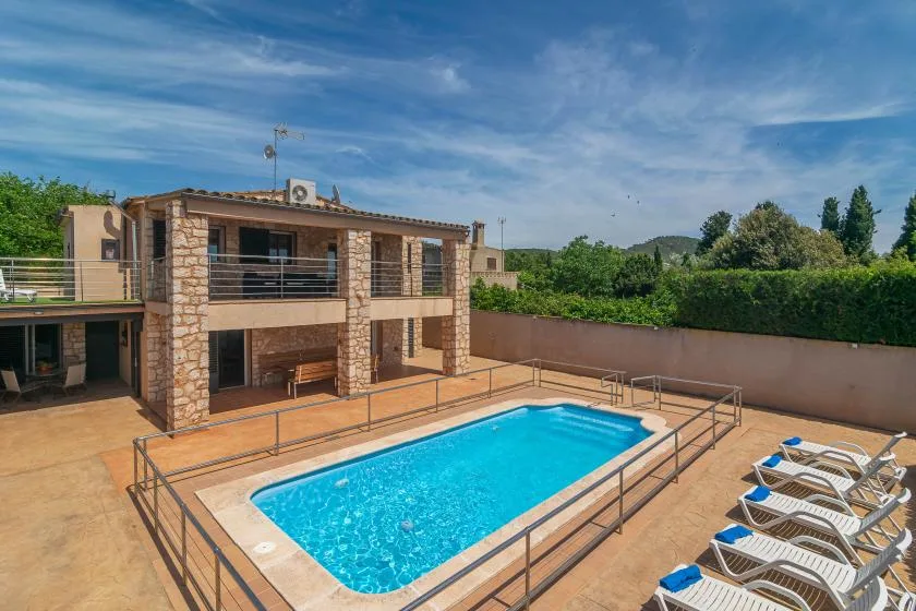 A swimming pool sits beside a two-story house and rows of lounge chairs.