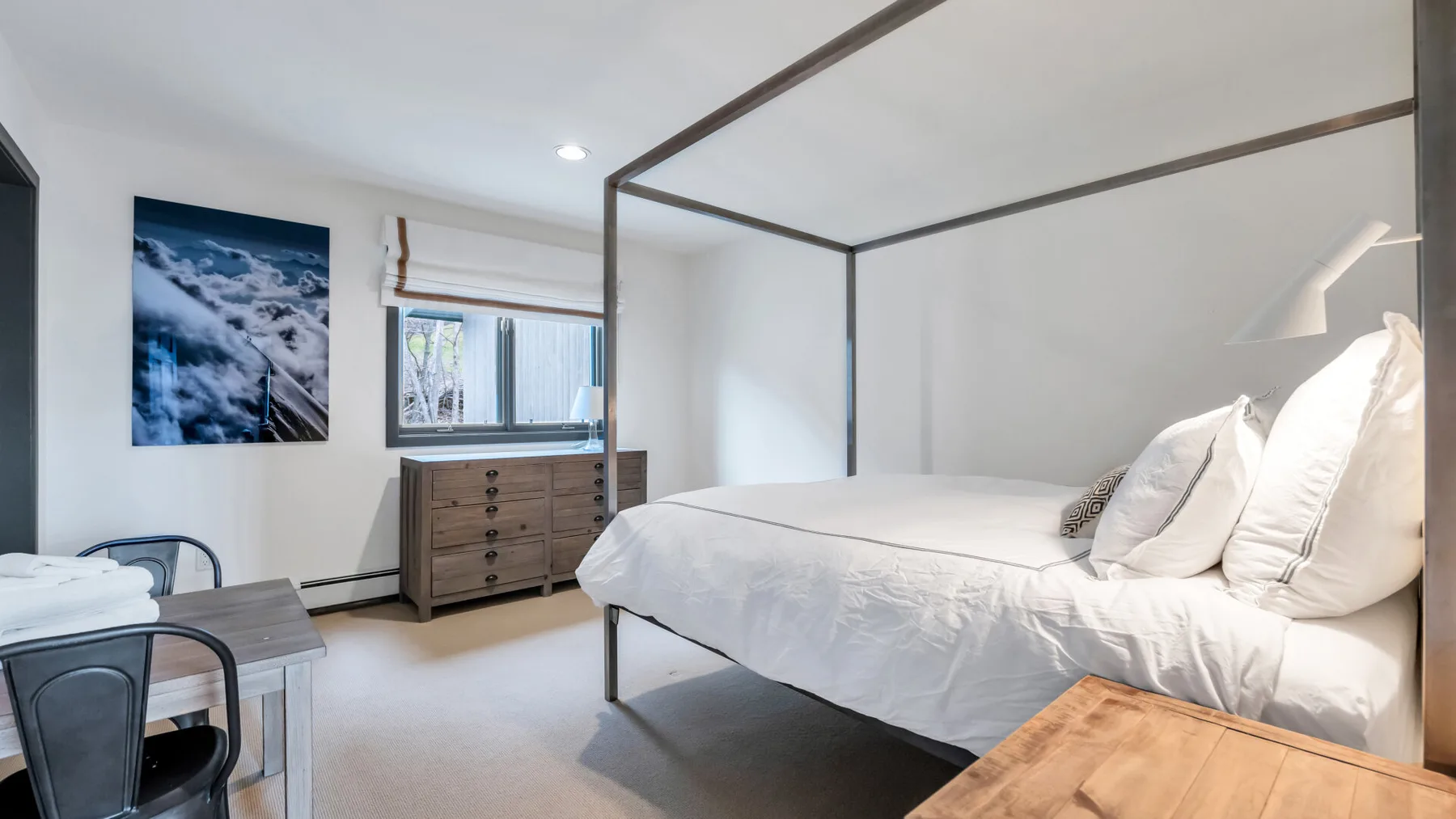 Un dormitorio luminoso con una cama blanca, una cómoda de madera y arte en la pared.