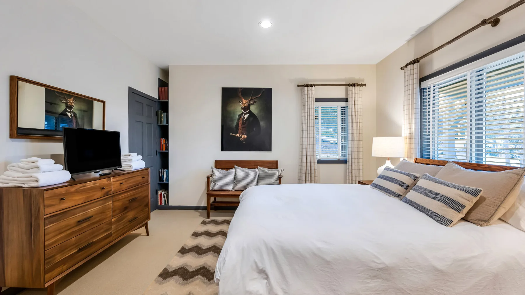 Un dormitorio luminoso con una cama grande, una cómoda de madera y ventanas con persianas.