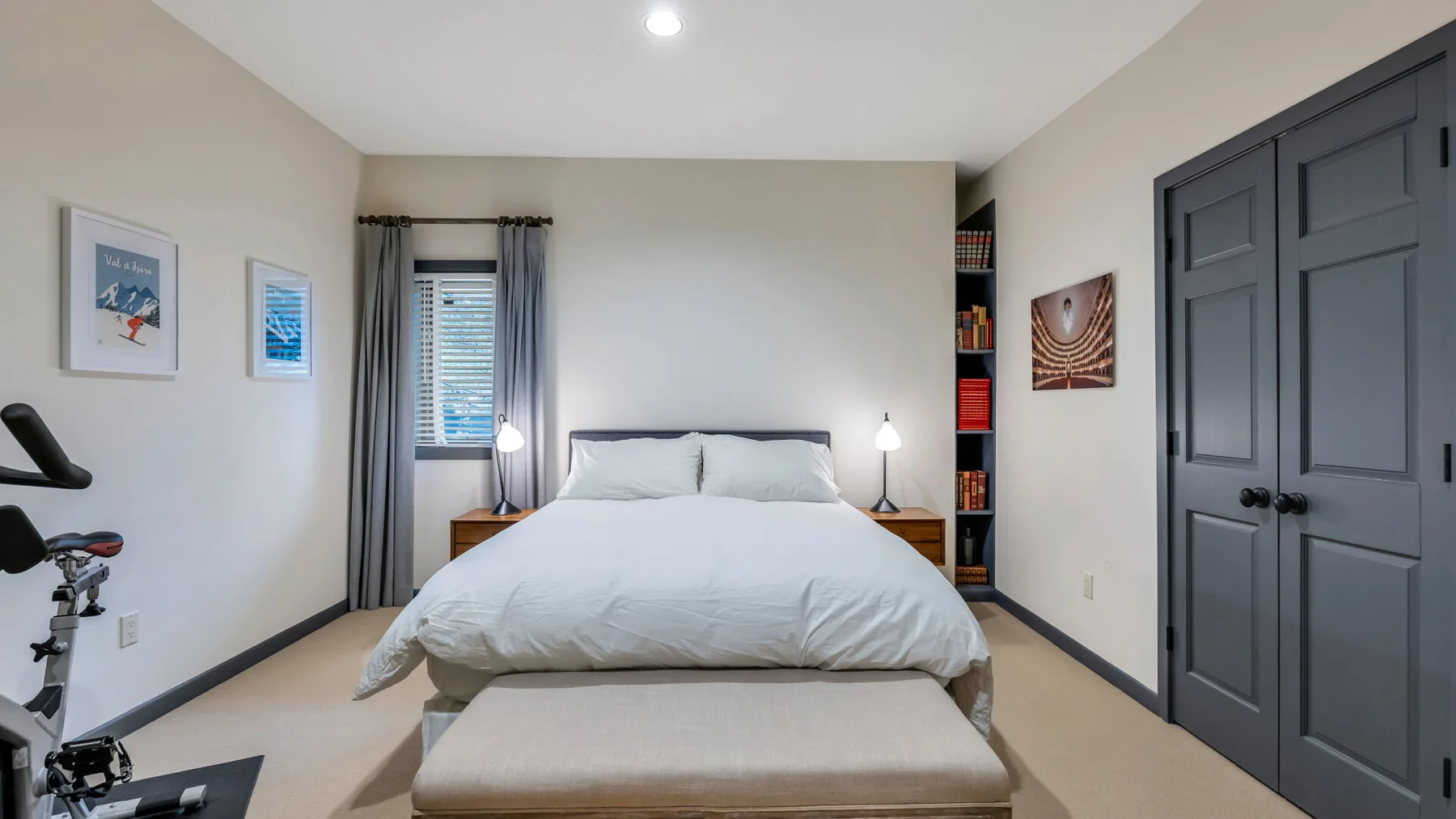 Un dormitorio con una cama bien arreglada, un banco y ventanas con cortinas.