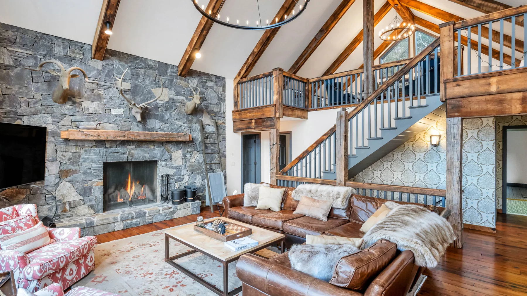 A rustic living room with a stone fireplace, seating, and a wooden staircase.