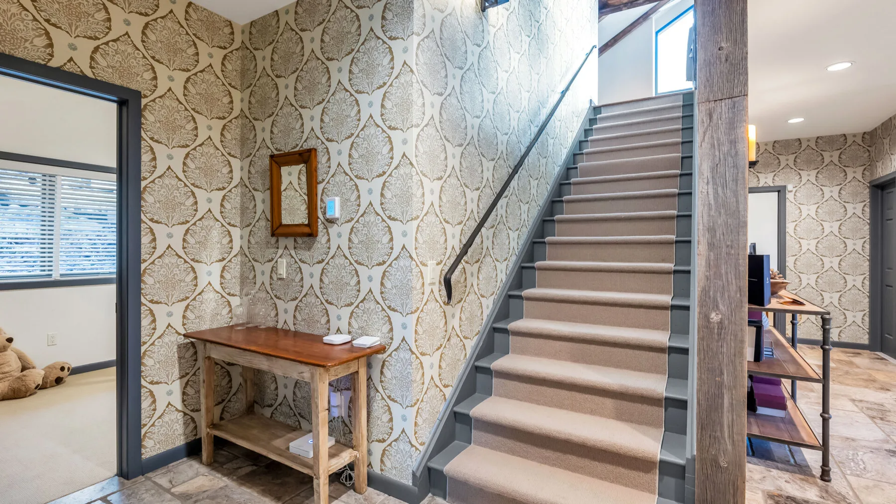 Una escalera alfombrada sube junto a un papel pintado estampado y una pequeña mesa de madera.