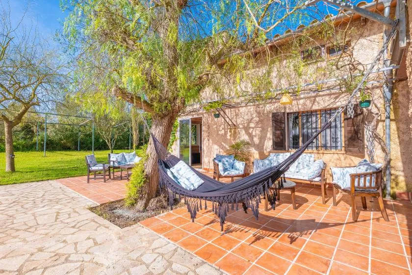 Un patio soleado con una hamaca, asientos al aire libre y un árbol frondoso junto a la casa.