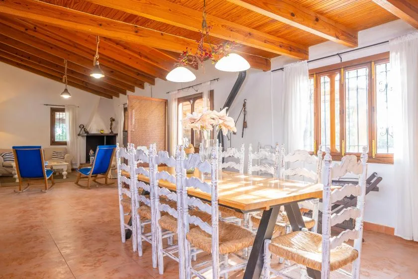 Un comedor con una mesa larga y sillas blancas bajo vigas de madera vistas.