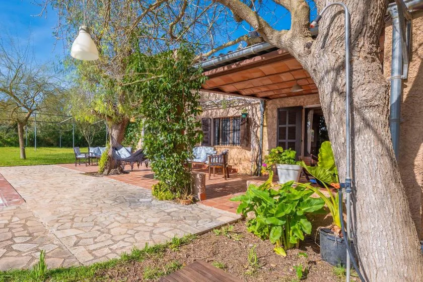Un patio de piedra junto a una casa, con asientos al aire libre y plantas frondosas.