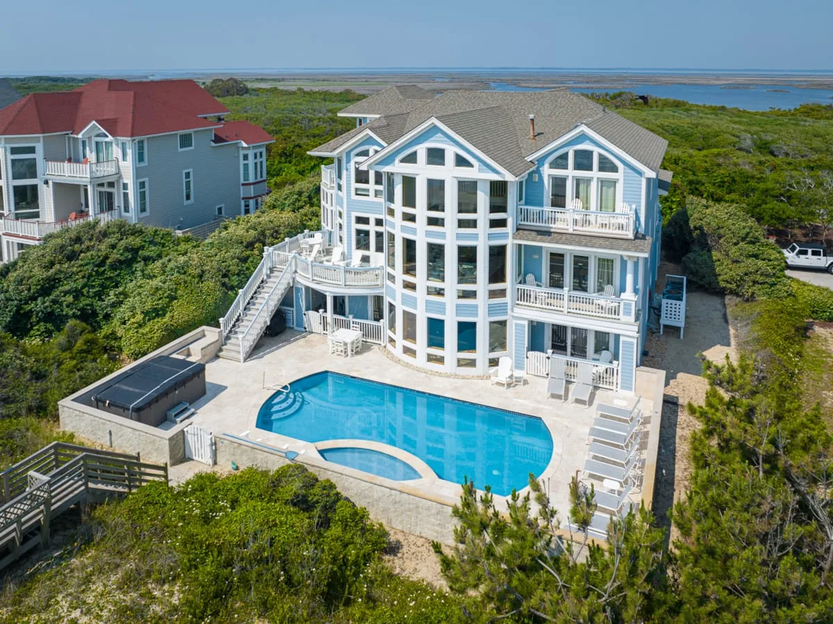 A large coastal house overlooks surrounding greenery, with a swimming pool in front.