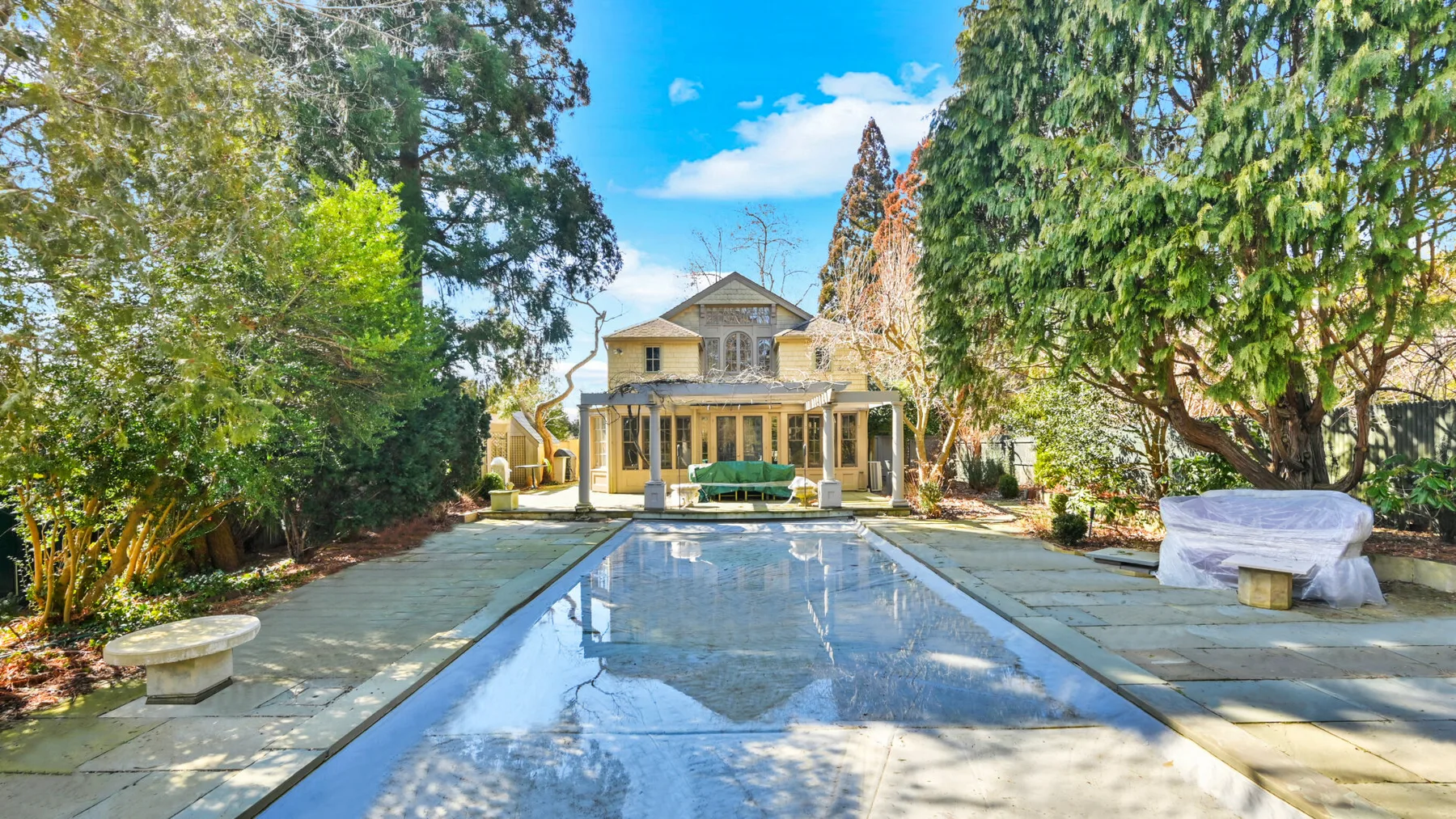 A swimming pool extends toward a house, with paved paths and trees around it.