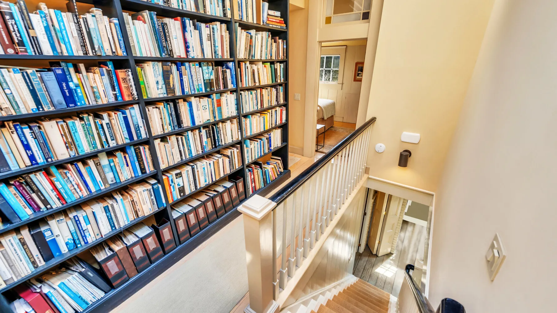 A tall bookshelf lines a staircase landing beside a wooden railing.