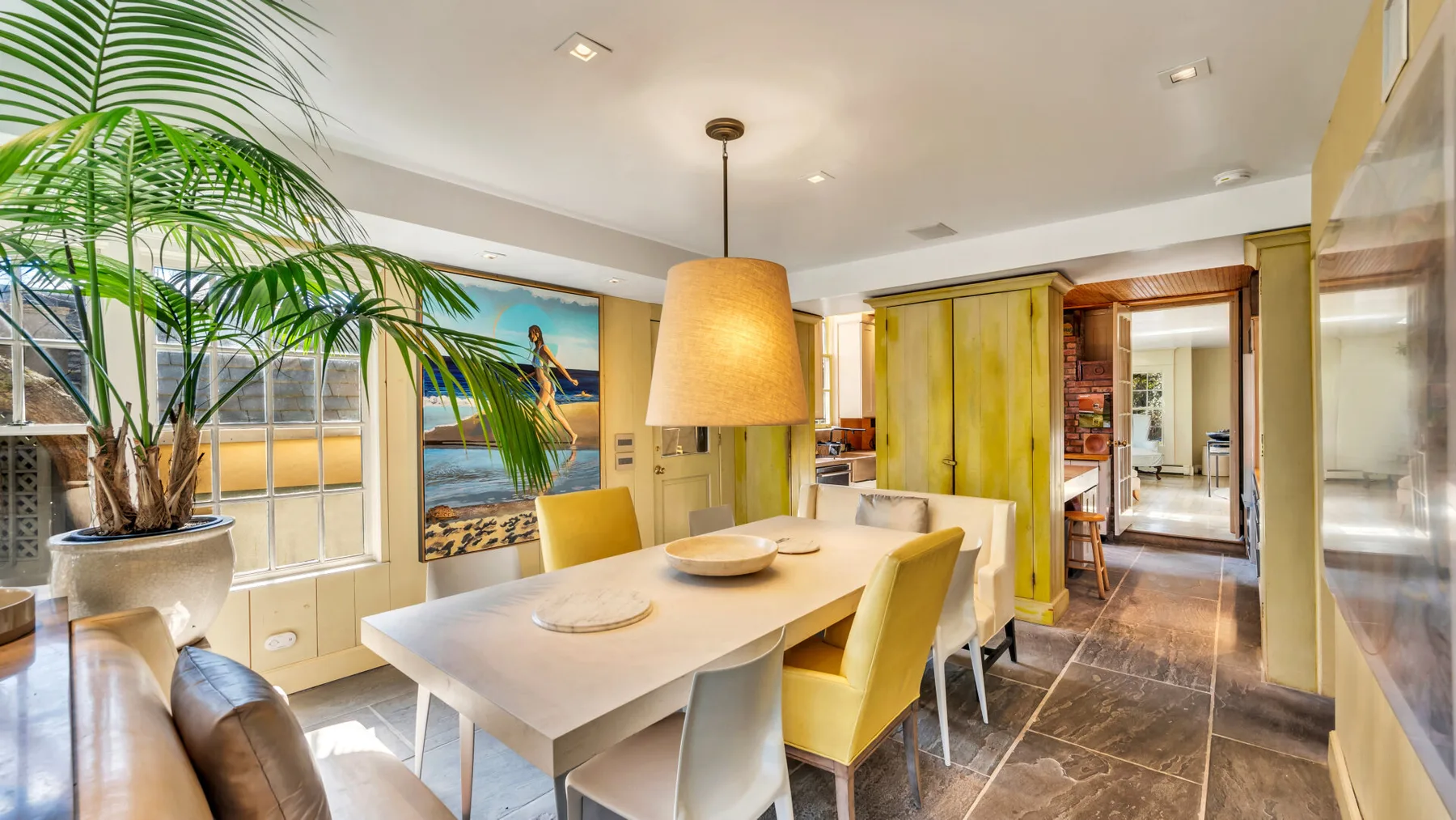 A dining area with a table and chairs beside large windows and leafy plants.
