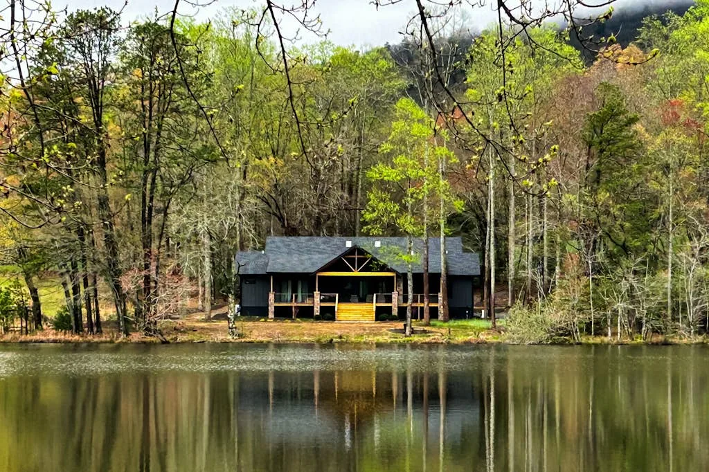 A dark-roofed cabin stands among trees beside a calm pond.