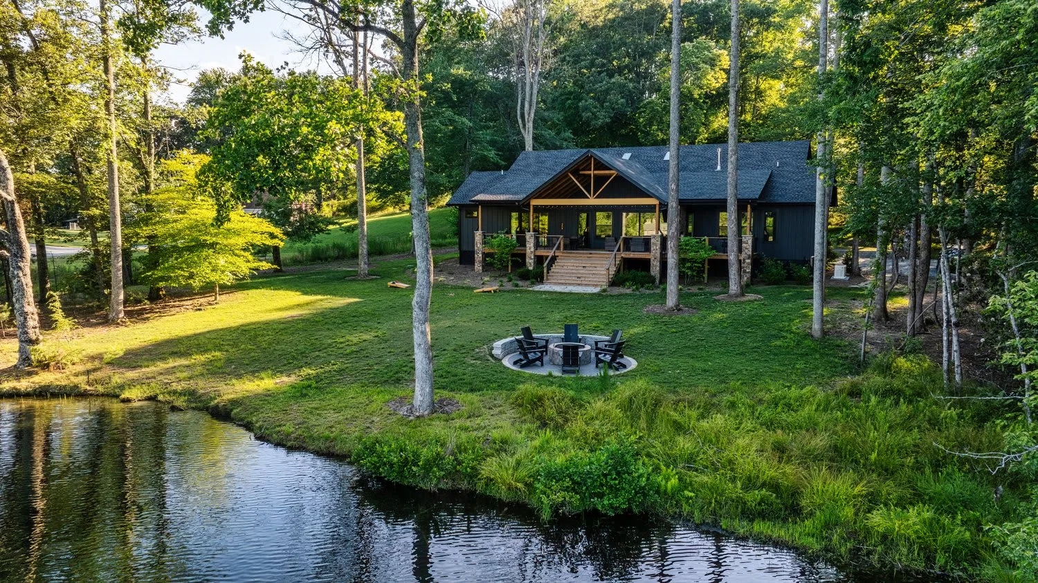A cabin sits among trees beside a grassy lawn and a small pond.