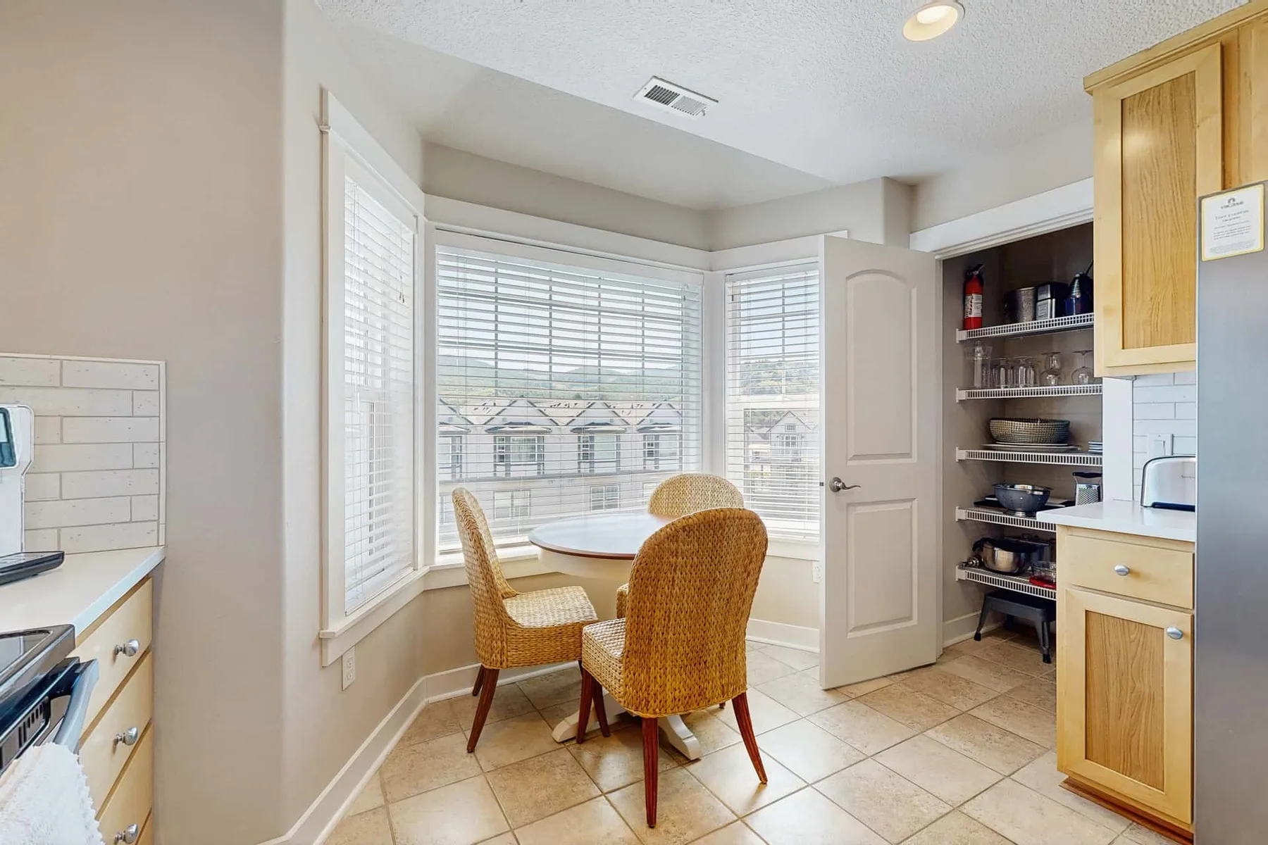 A bright kitchen dining area with a round table and wicker seating beside large windows.