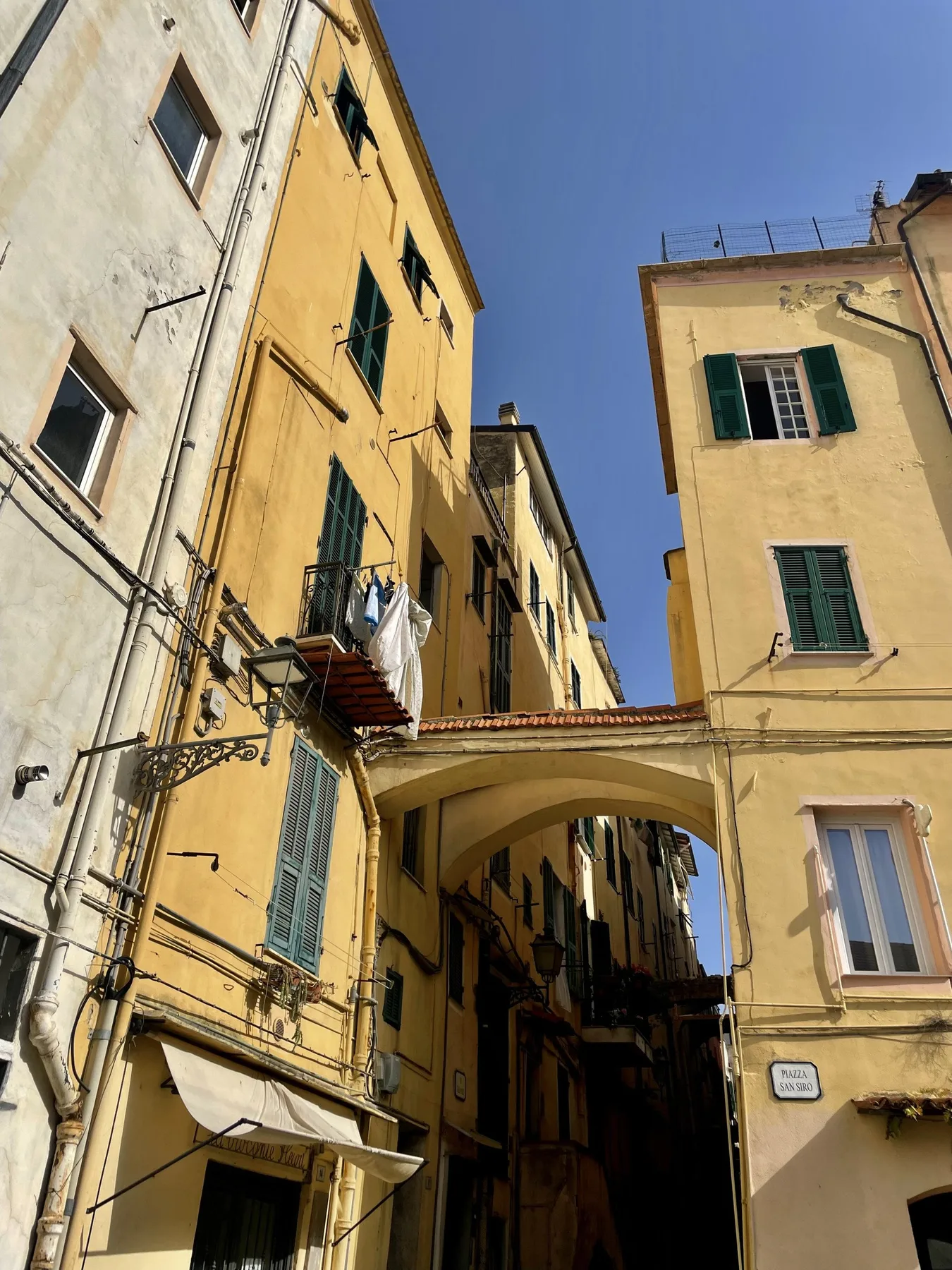A narrow passage runs between tall, sunlit buildings beneath a clear blue sky.