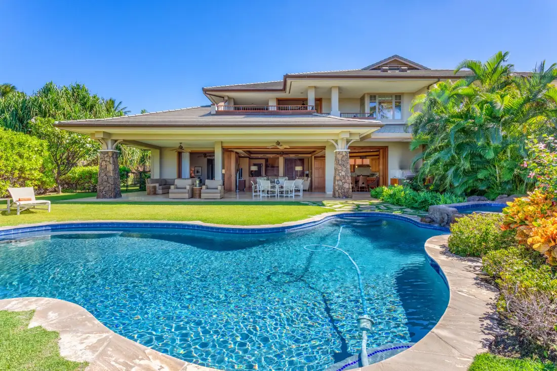 A swimming pool sits in front of a house with a covered patio and tropical landscaping.