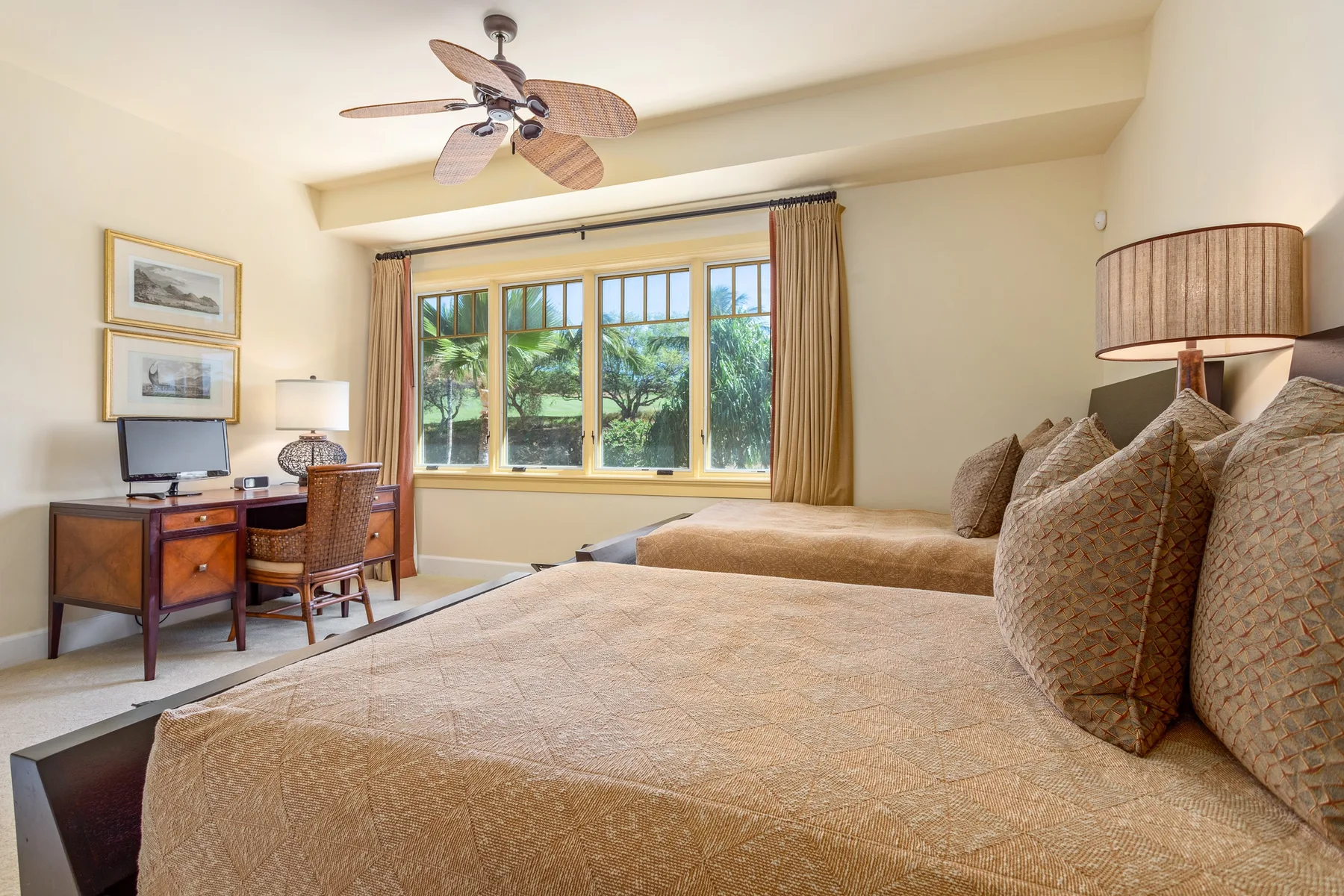 A bedroom with two beds, a desk, and windows overlooking greenery.