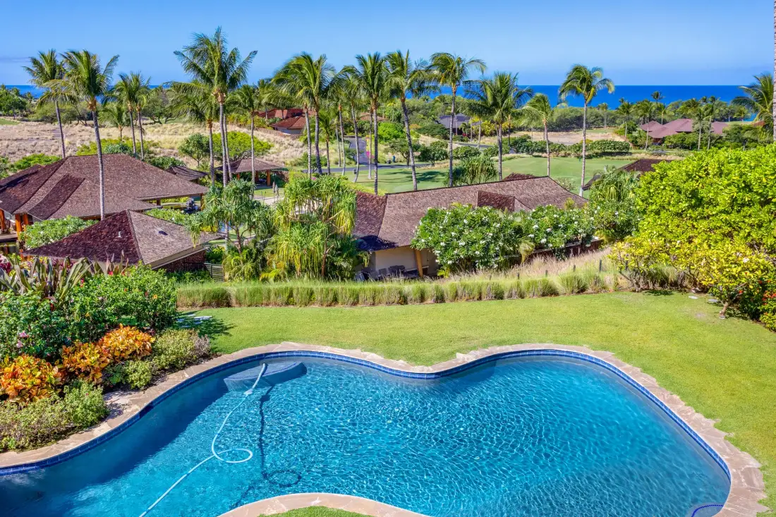 A swimming pool overlooks landscaped grounds, palm trees, and the ocean.