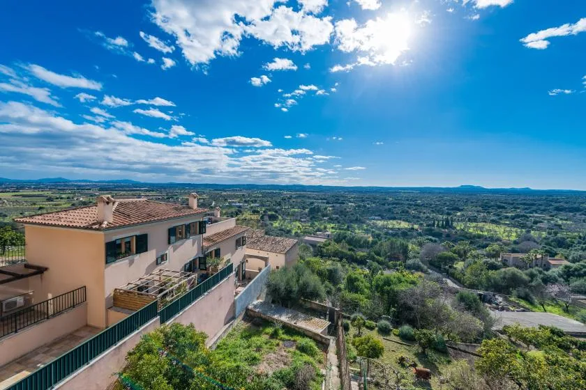 A hillside residential building overlooks a broad green landscape beneath a bright blue sky.