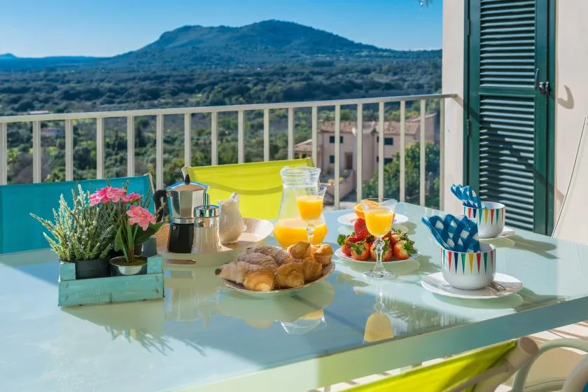 A balcony table is set with breakfast and flowers, overlooking green hills.