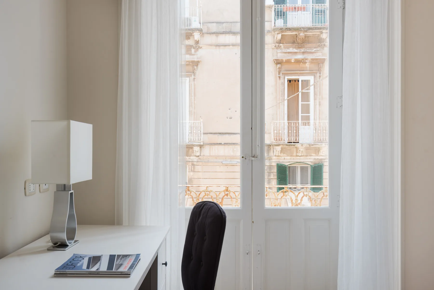Una habitación luminosa con un escritorio junto a puertas acristaladas y cortinas translúcidas.