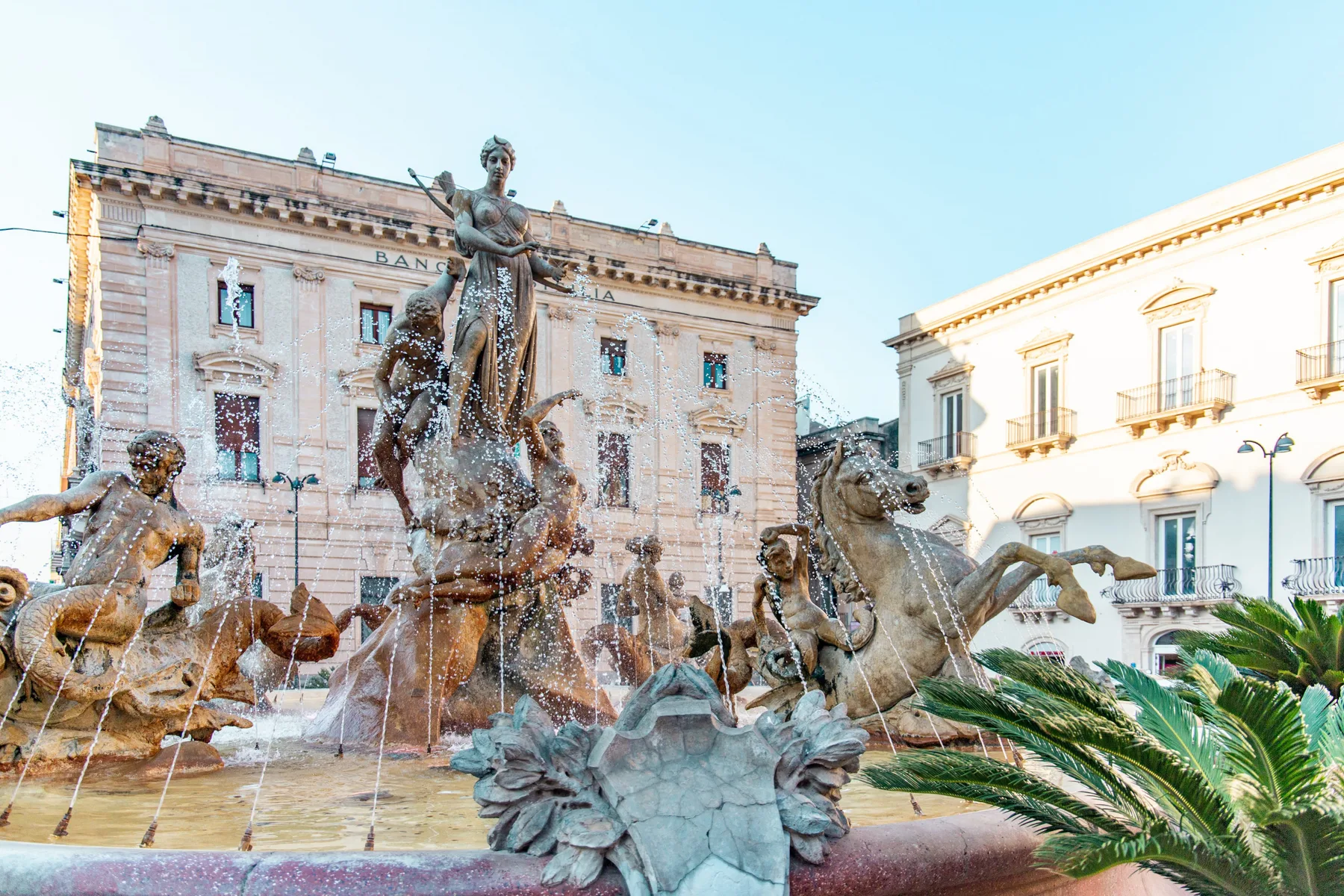 Una gran fuente escultórica se encuentra en una plaza entre edificios de aspecto histórico.