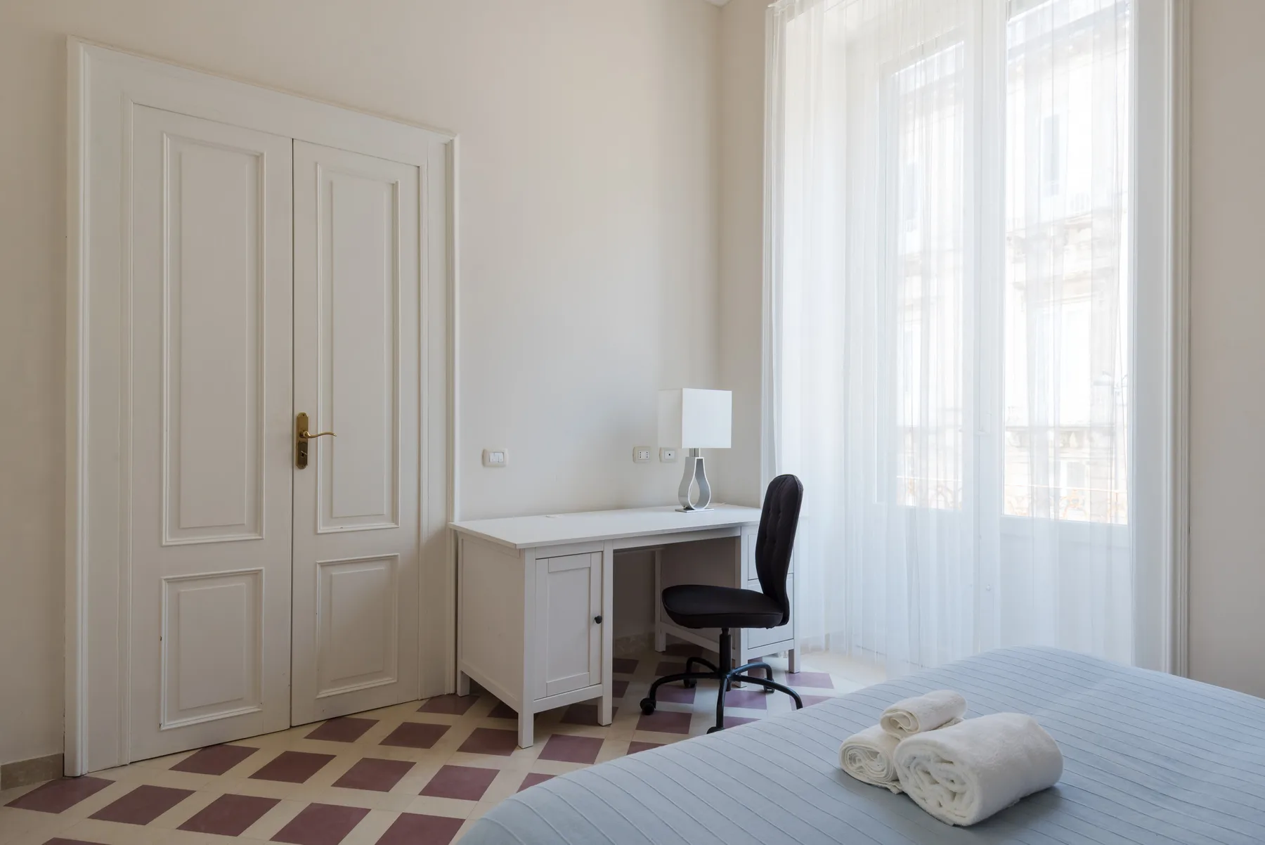 Un dormitorio luminoso con un escritorio junto a una ventana grande con cortinas translúcidas.