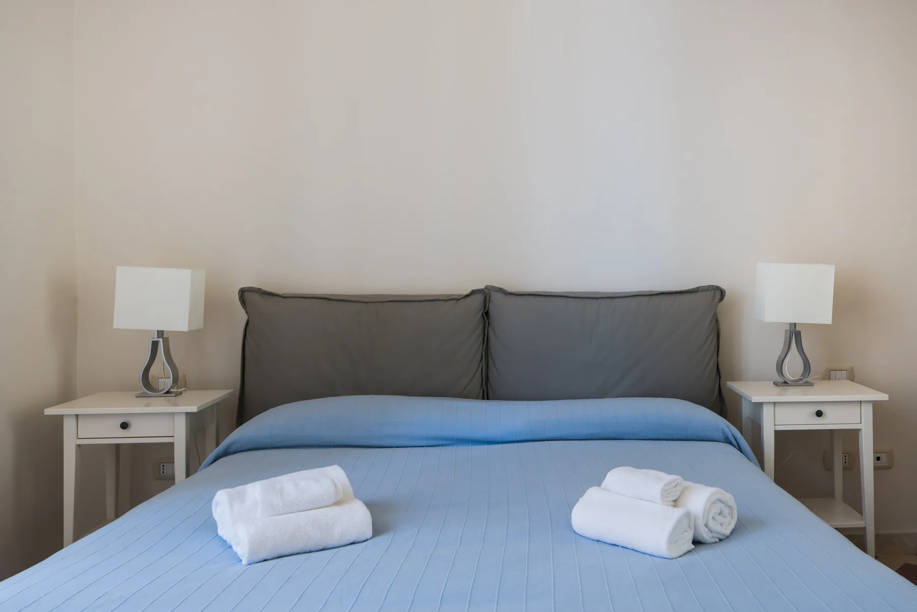 Un dormitorio con una cama azul, almohadas grises y mesitas de noche a juego.